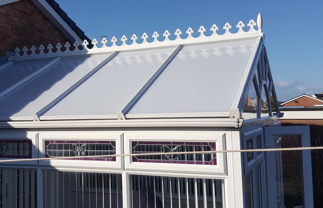 Interior view of a conservatory roof with triangular glass panels and a central hub fitted with clamps during repair work, with a ladder visible outside the window.