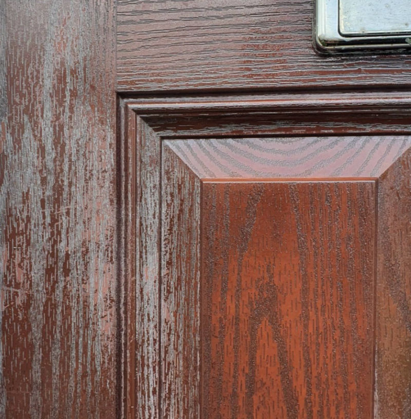 The image shows a section of a rosewood composite door with the color being restored. The door features a detailed wood grain pattern with a reddish-brown hue. The image highlights an area where the color appears to be in the process of being restored, as evidenced by the contrast between the more vibrant, restored sections and the duller, worn sections. This is interesting and relevant as it demonstrates the process and effect of restoring the color and finish of a composite door, which can be useful for understanding maintenance and care for such materials