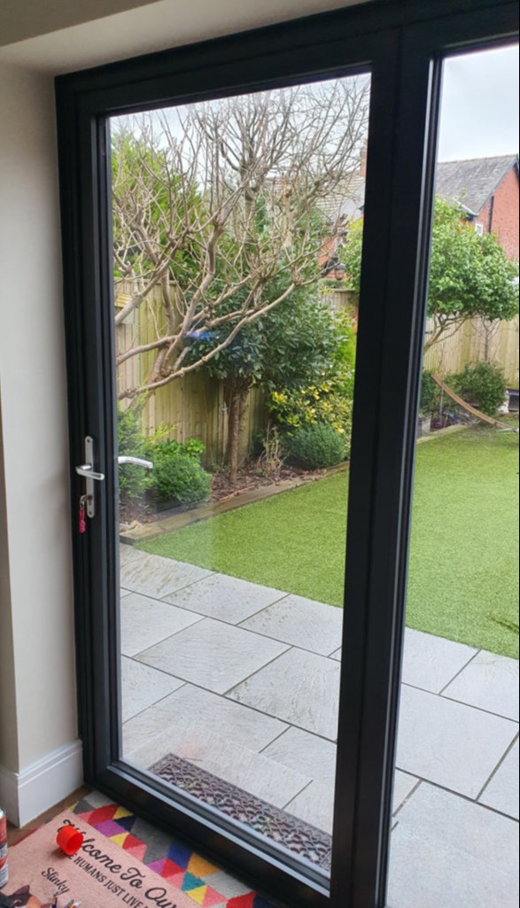The image shows a view through a large glass bi-folding door looking out onto a backyard garden. The garden features a well-maintained lawn, a paved patio area, and various trees and shrubs along the fence. There is a colorful doormat inside the house with text that reads: "Welcome To Our Home. Humans Just Live Here." The image captures a serene and tidy outdoor space, providing a glimpse into a well-kept garden area.