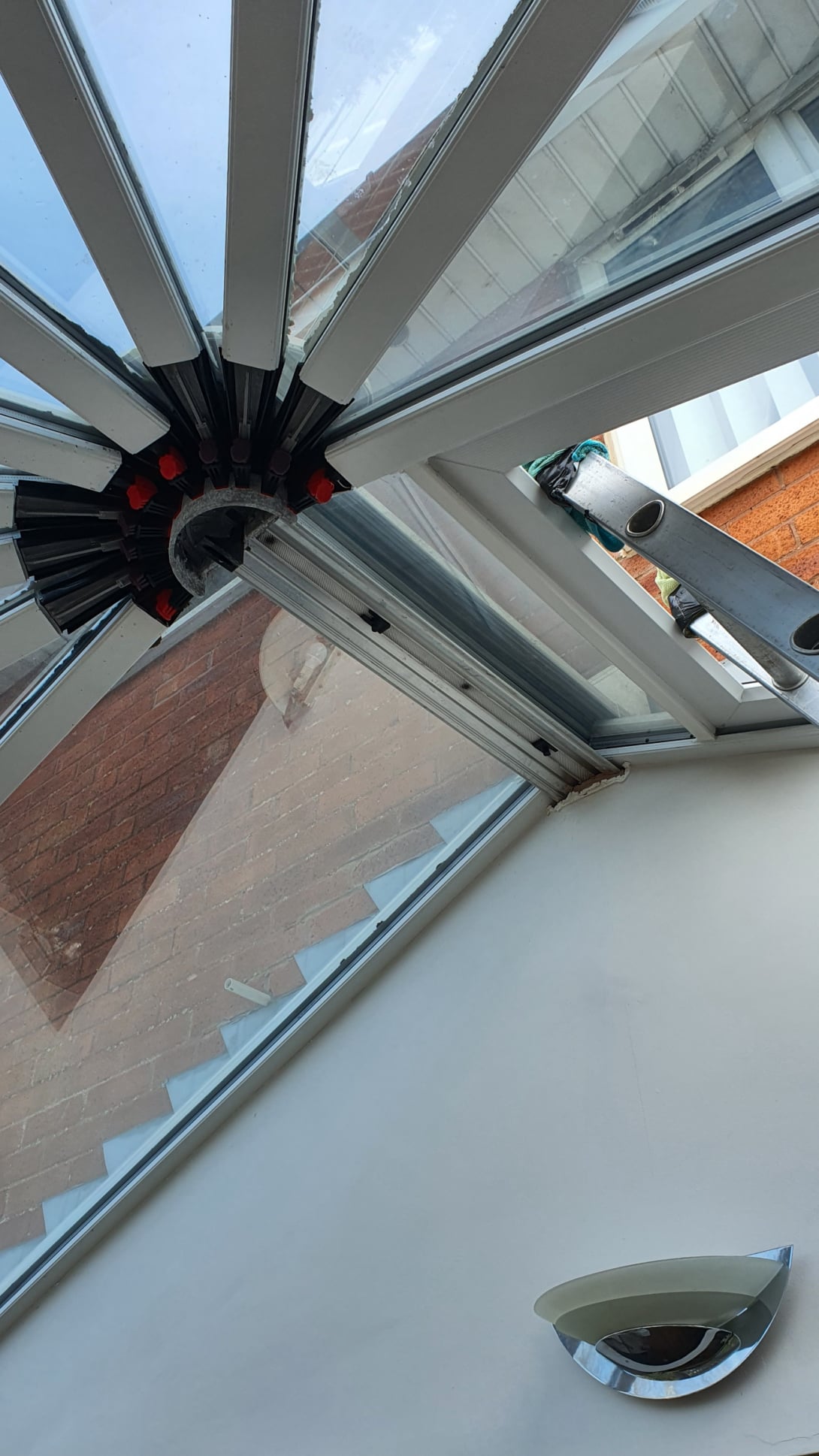 Interior view of a conservatory roof with triangular glass panels and a central hub fitted with clamps during repair work, with a ladder visible outside the window.