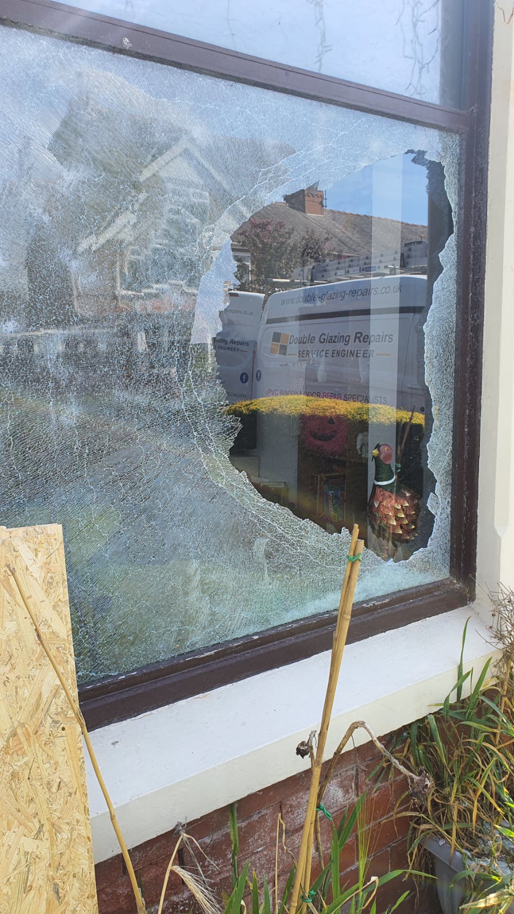 “Broken double‑glazed window with a large hole and shattered glass, with a Double Glazing Repairs service van visible outside.