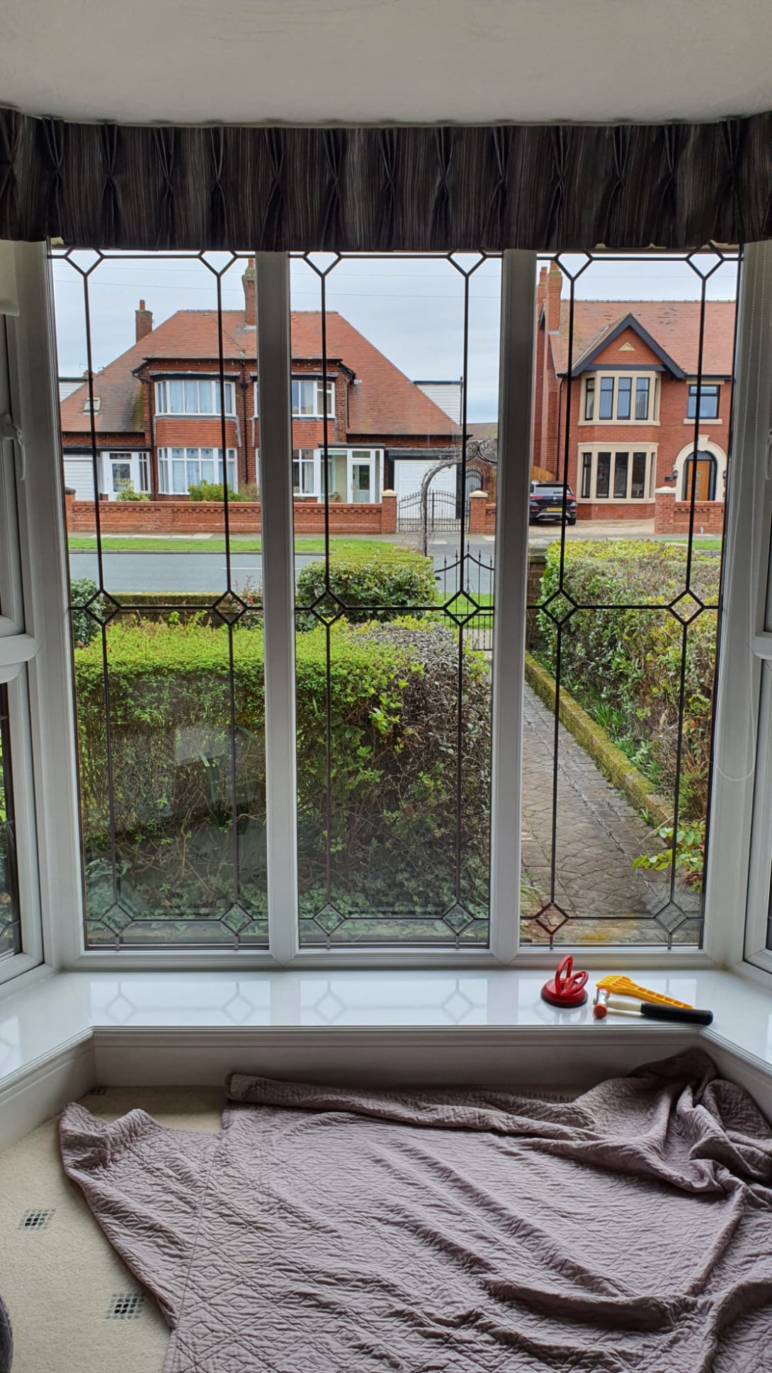 Bay window with decorative leaded glass being prepared for replacement, with tools on the sill and a residential street visible outside.