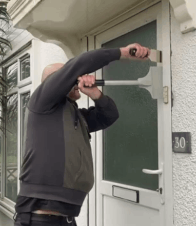 The image shows a person using a door spreading tool to open a locked uPVC door. The tool is being inserted between the door and the frame, likely to manipulate the locking mechanism or create enough space to unlock the door. This image is relevant as it demonstrates a method for gaining entry through a locked uPVC door without causing significant damage.